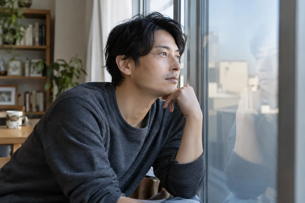 窓に映る自分を静かに見ながら考える男性